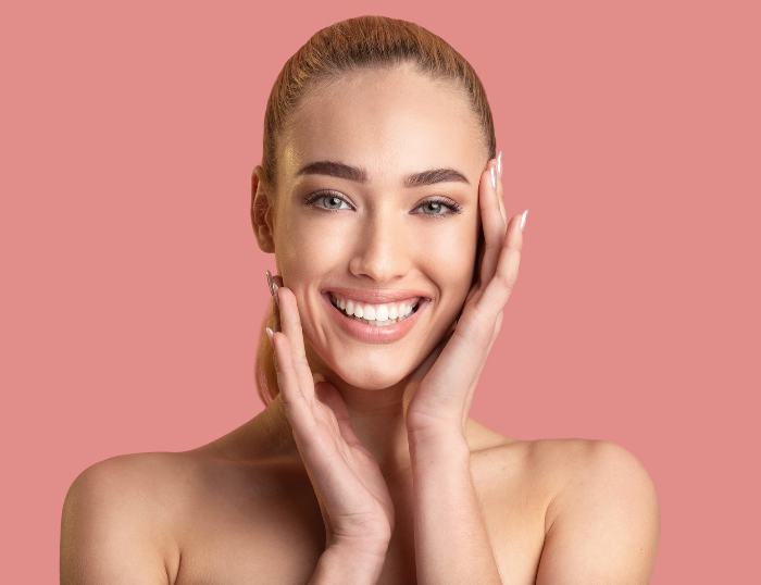 Junge Frau mit strahlender Haut, die beide Hände sanft ans Gesicht legt, vor rosa Hintergrund.