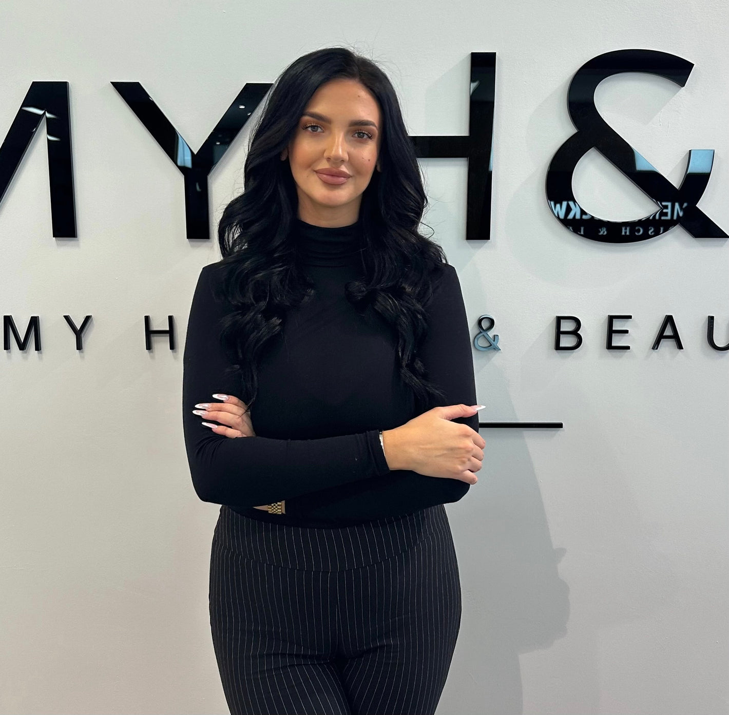 Mitarbeiterin von MYH&B mit langen schwarzen Haaren vor Logo-Wand – selbstbewusste Ausstrahlung