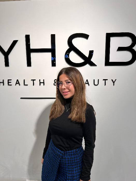 Porträt einer MYH&B Mitarbeiterin mit langen Haaren und Brille vor der Logo-Wand